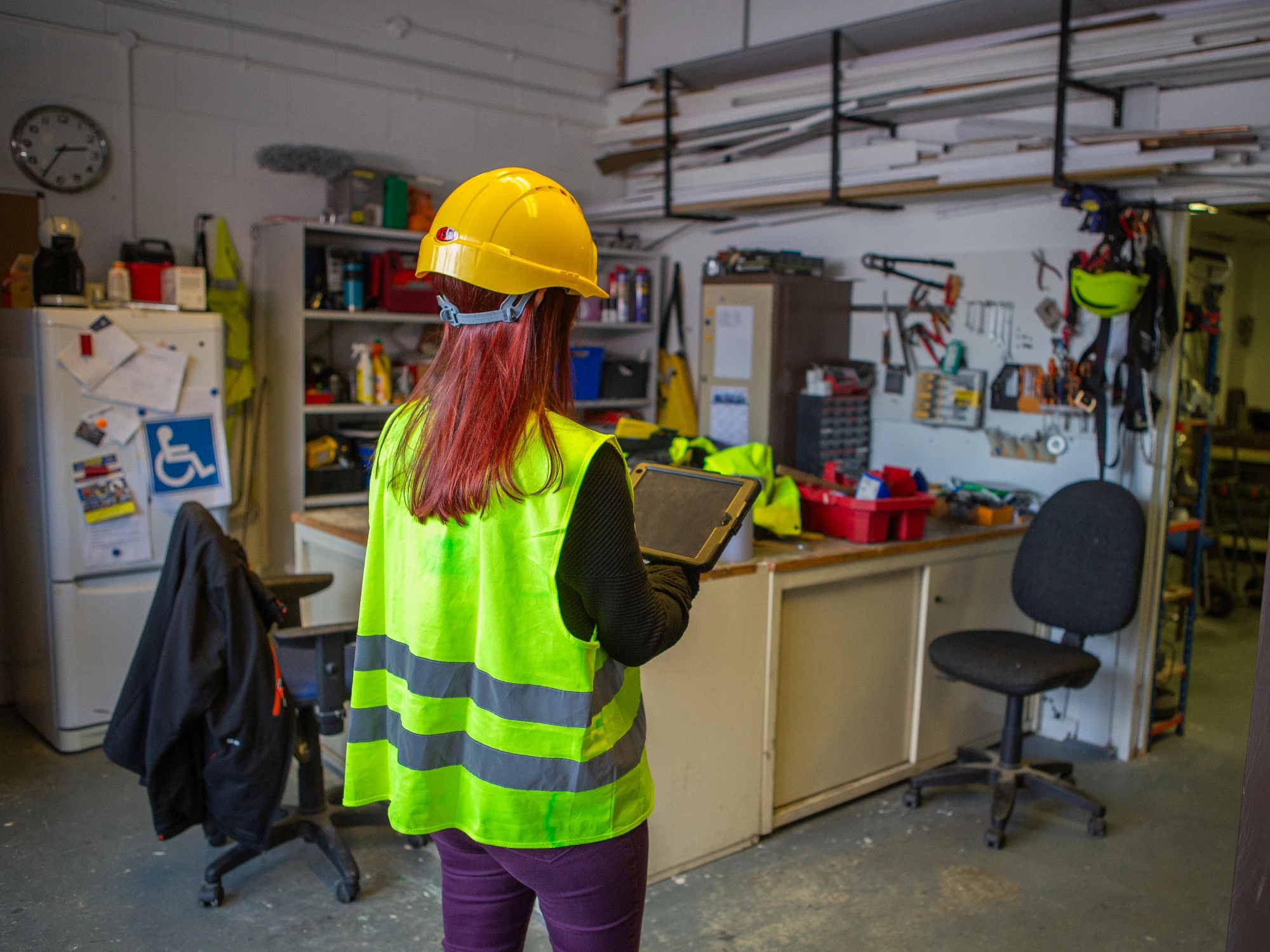A woman in a yellow hardhat and hi-vis uses a tablet to check safety information. 